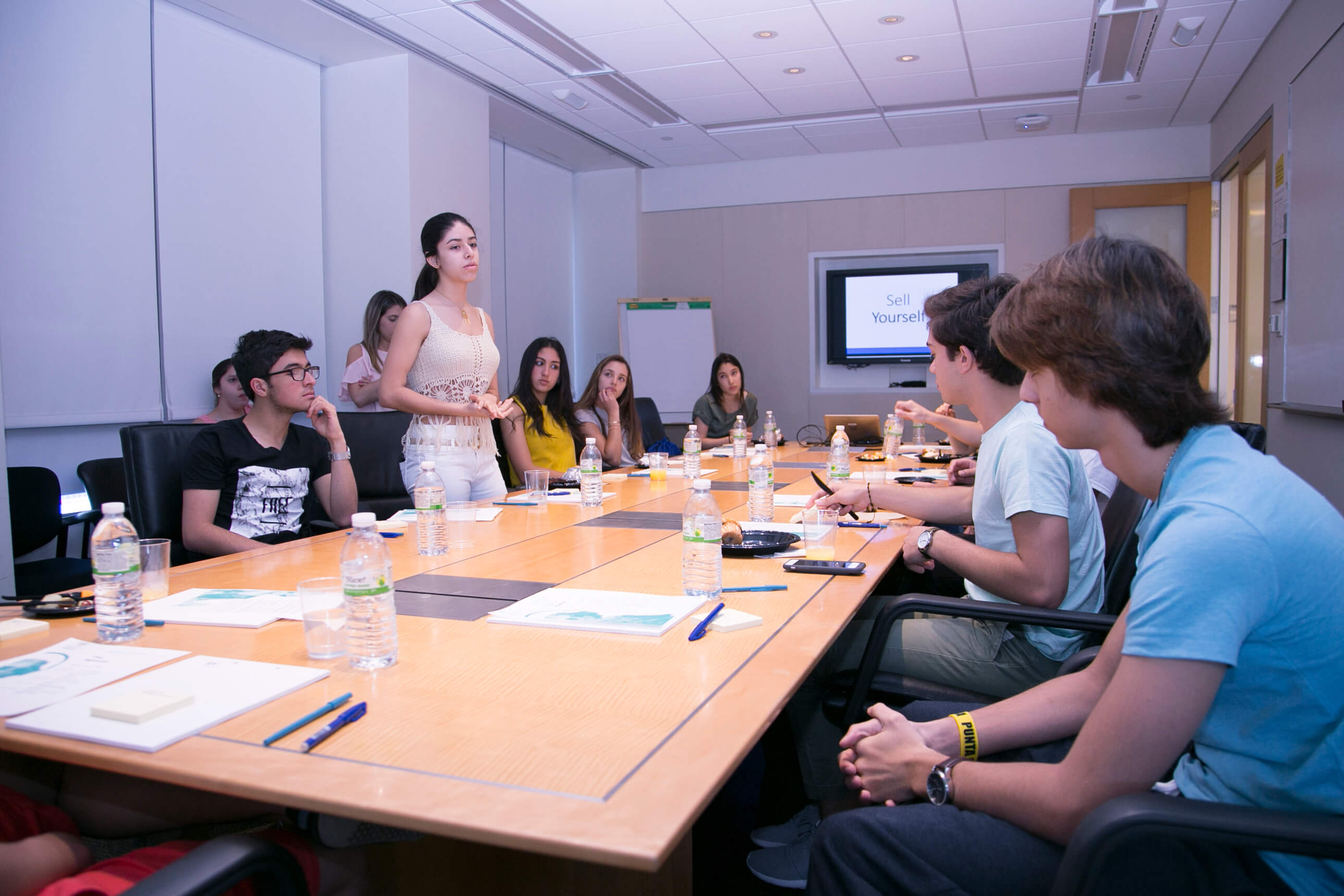 A Visionarium student standing to address the group across a boardroom table during the public-speaking workshop.